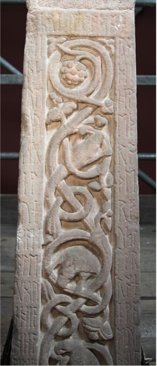 A close-up of inhabited vine-scroll on the Ruthwell Cross, carved in relief on pink-grey sandstone. Old English inscriptions in runes are engraved in the borders.