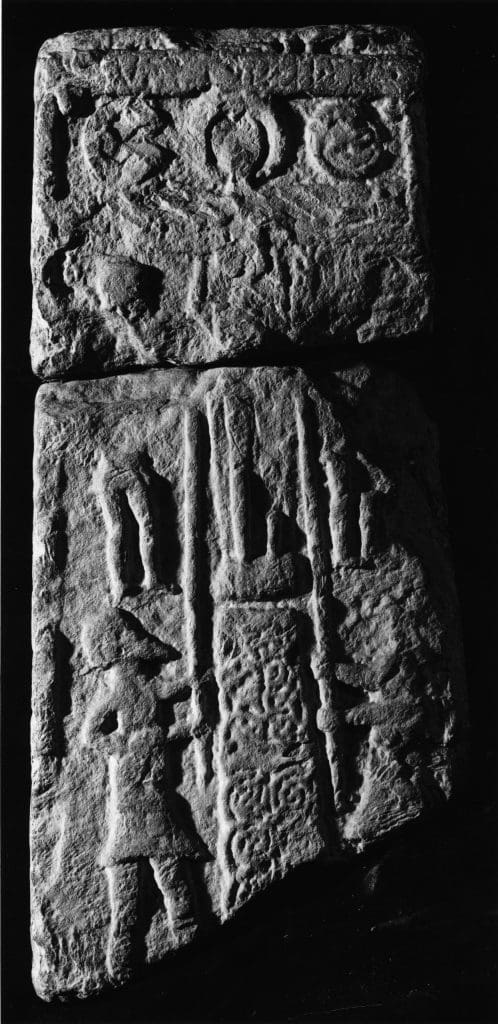 A black and white photograph of a broad face of a cross-shaft fragment. The engravings depict the crucifixion, including Stephaton and Longinus with the spear and sponge. Above the crucifixion scene is a now-illegible inscription.