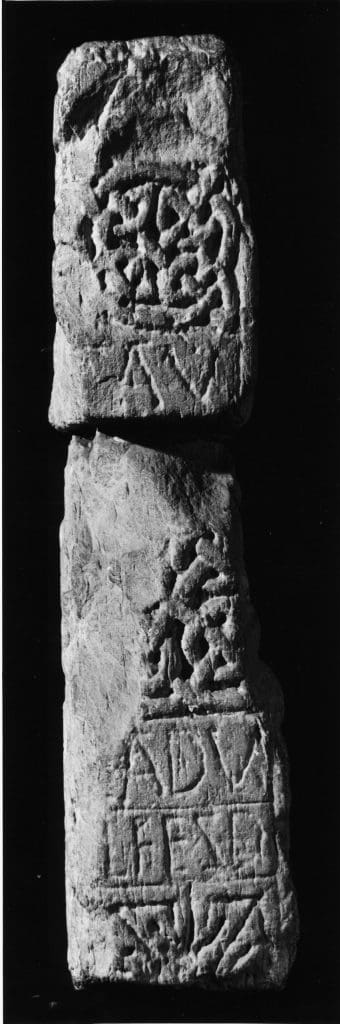 A black and white photograph of a narrow face of a cross-shaft fragment. Three panels of interlace divided horizontally by two inscriptions in Latin capitals.