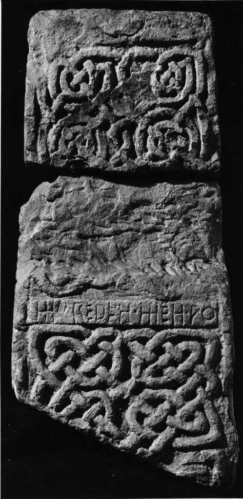 A black and white photograph of a broad face of a cross-shaft fragment. Two panels of interlace divided horizontally by an inscription in a mix of Latin capitals and runes. 