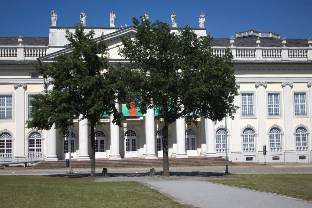 Two oak trees paired with two basalt columns, located in front of a white columned building.
