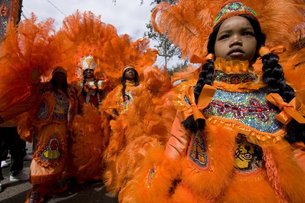 Participants in Black Masking Indians festivities pose for a photograph: a young child gazes solemnly out in the right foreground, while three adult participants stand together in the left midground, all wearing florescent orange feathered costumes with beadwork.