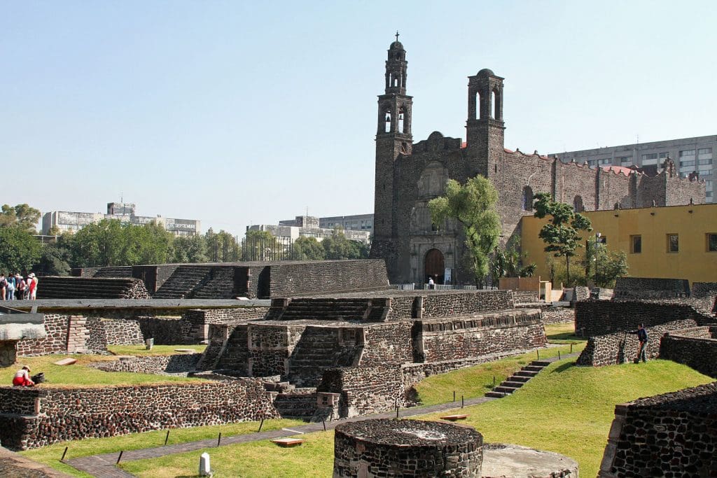 View across an archeological site with ruins of a Mexica temple with a church fabricated from the same stones in the background. 