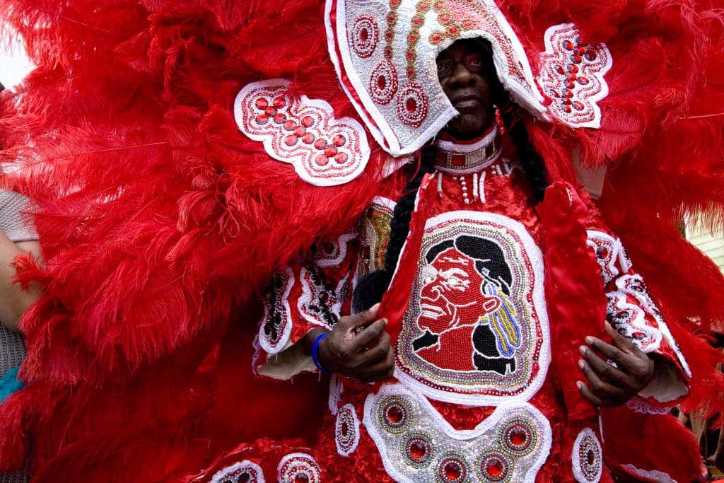 Participant in the Black Masking Indians festivities poses for a photograph: the performer displays his elaborate costume beadwork with the bust of a longhaired man; his red feathered costume fills the frame.