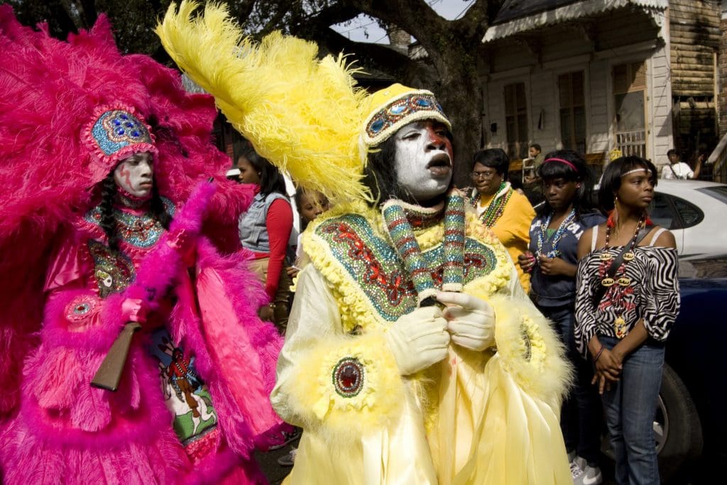 Black Masking Indian procession, with two costumed participants on the left foreground, and street-side viewers grouped around cars and in front of a white house on the right. 