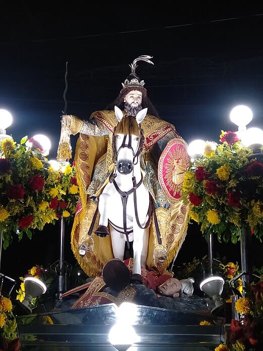 Statue of a knight with long hair on horseback (Santiago Matamoros), trampling severed heads, positioned on top of the silver hood of a car and surrounded by flowers and bright lights at night. 