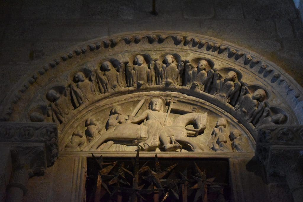 Relief carving in stone of a knight on horseback with a sword and cross-topped banner placed centrally in an arch over a bronze door. 