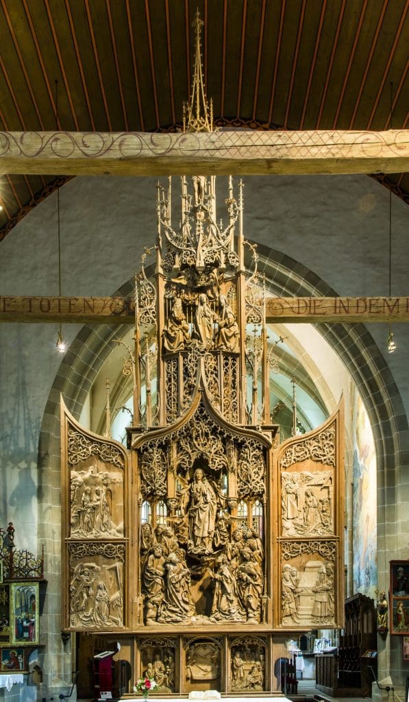 Photograph of a large, ornately carved wooden altarpiece with open, hinged doors in the center that reveal an image of a woman ascending up with a group of people watching from below.
