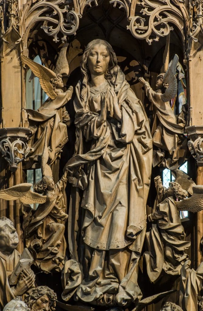 A close-up photograph of an ornately carved wooden altarpiece showing an image of a woman with her hands in prayer wearing voluminous robes.