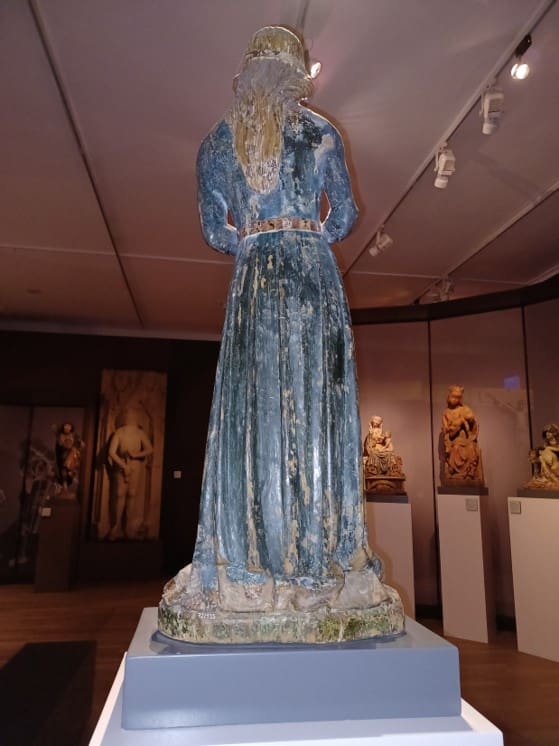 Photograph of the back of a sculpture of a standing woman with long hair wearing a floor-length gown.
