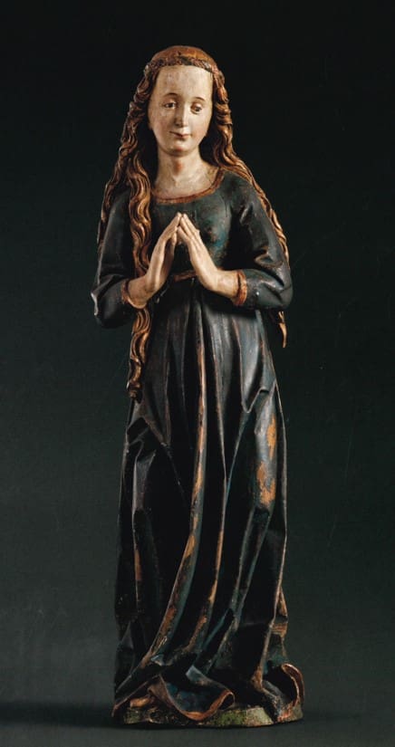 Photograph of a sculpture of a woman with long, blond hair with her hands in prayer. She wears a simple floor-length dress.