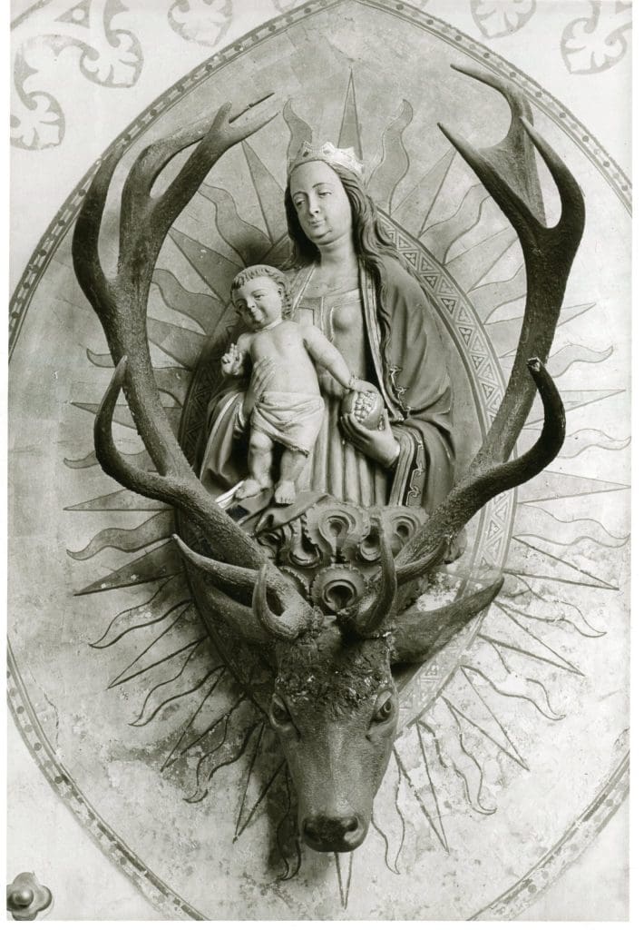 A photograph of a sculpture of a woman wearing a crown and holding a small child. The two figures rest on top of the head of a stag, whose antlers curve up and around the figures.