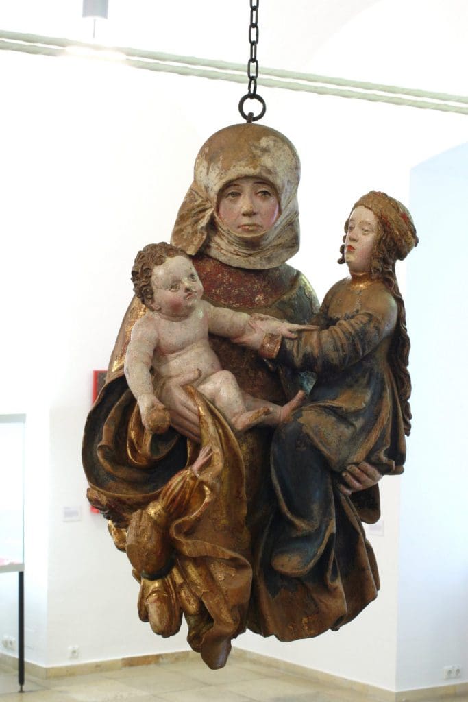 Photograph of a sculpture of an older woman holding a younger woman in her left hand and a baby in her right hand. The whole sculptural group hangs from a chain.