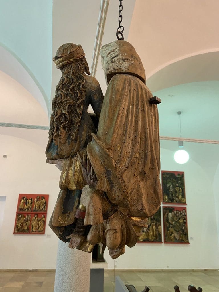 A side-view photo of the previous image, showing the back of an older woman wearing heavy robes holding a younger woman. The younger woman has long hair flowing down her back.