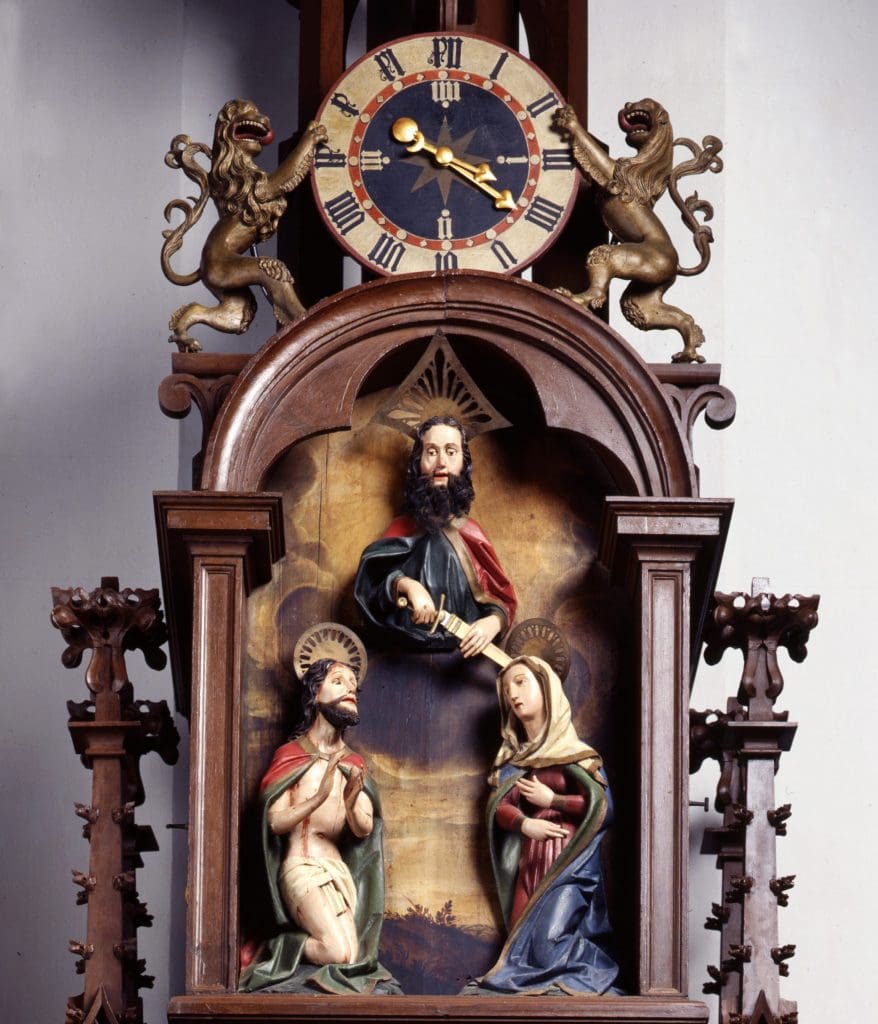 Photograph of a clock face above a niche containing a sculpture of three figures--a man holding a sword floats above a kneeling man and a kneeling woman.