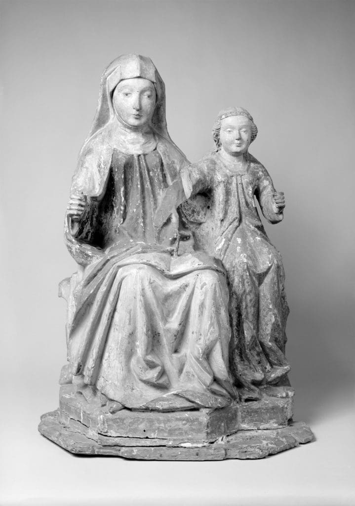 Black and white photograph of a sculpture of two female. The woman on the left is larger and wears robes and a headdress. The girl on the right is smaller and her hair is visible. 