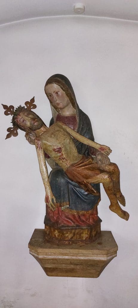 Photograph of a sculpture depicting a woman wearing a blue cloak holding a dead, male, emaciated body wearing a crown. 