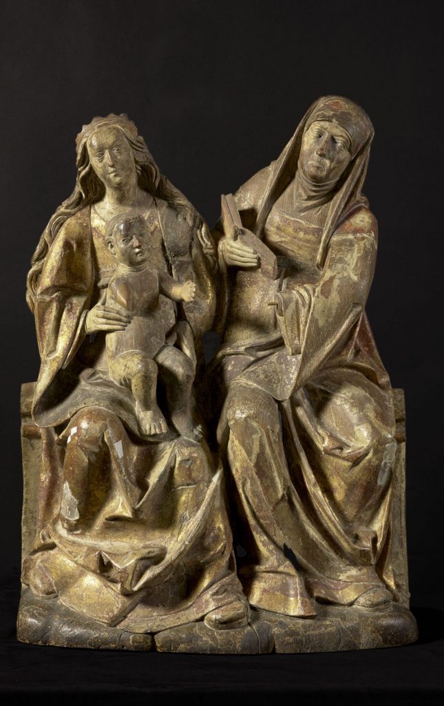 Photograph of a sculpture of three figures, includin an older woman with a book in her hands and a younger woman holding a child in her lap.