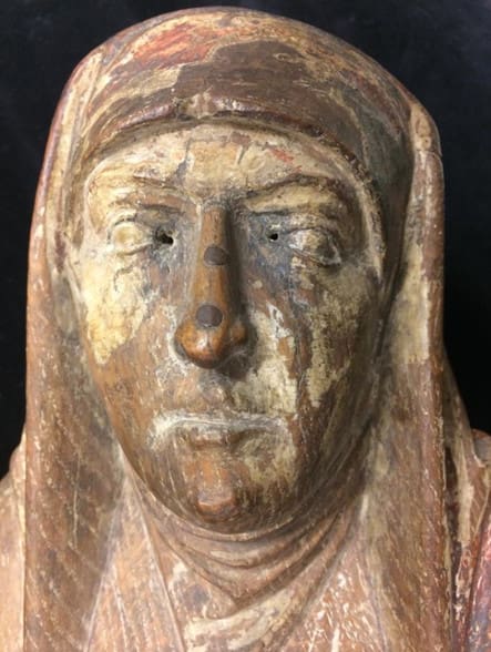 Close-up photograph of a wooden sculpture of a woman wearing a headdress, showing the holes in the inner corners of the eyes.
