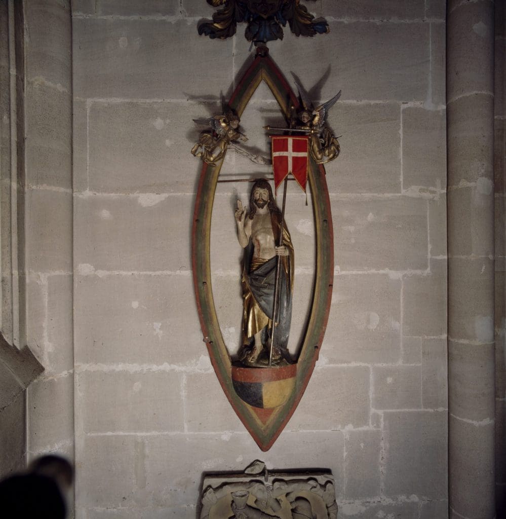 Photograph of a sculpture of a standing male figure within an almond-shaped frame. The man holds up his right hand and holds a flag in his left hand.
