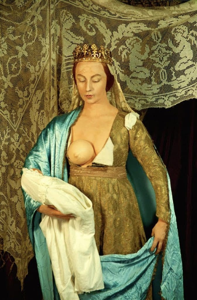 Photograph of the artist Cindy Sherman posing as the Virgin Mary before a curtain with angel motif, her open dress revealing a rubber breast and a doll in a white cloth on her right arm.