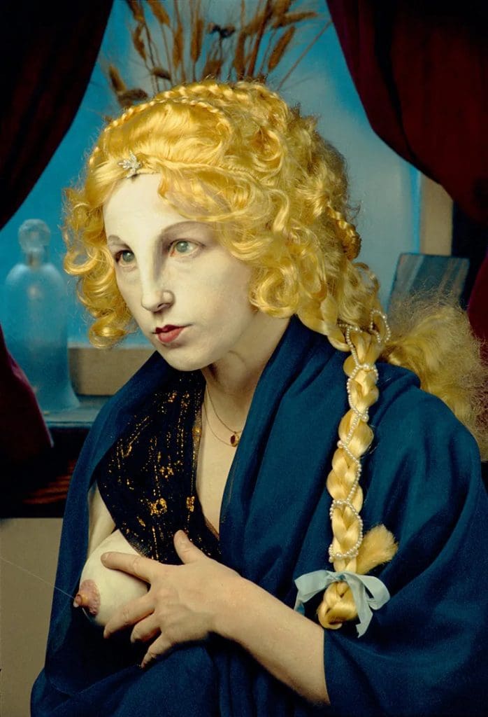 Photograph of the artist Cindy Sherman posing with a blonde wig and prosthetic breast, from which a drop of liquid dangles.